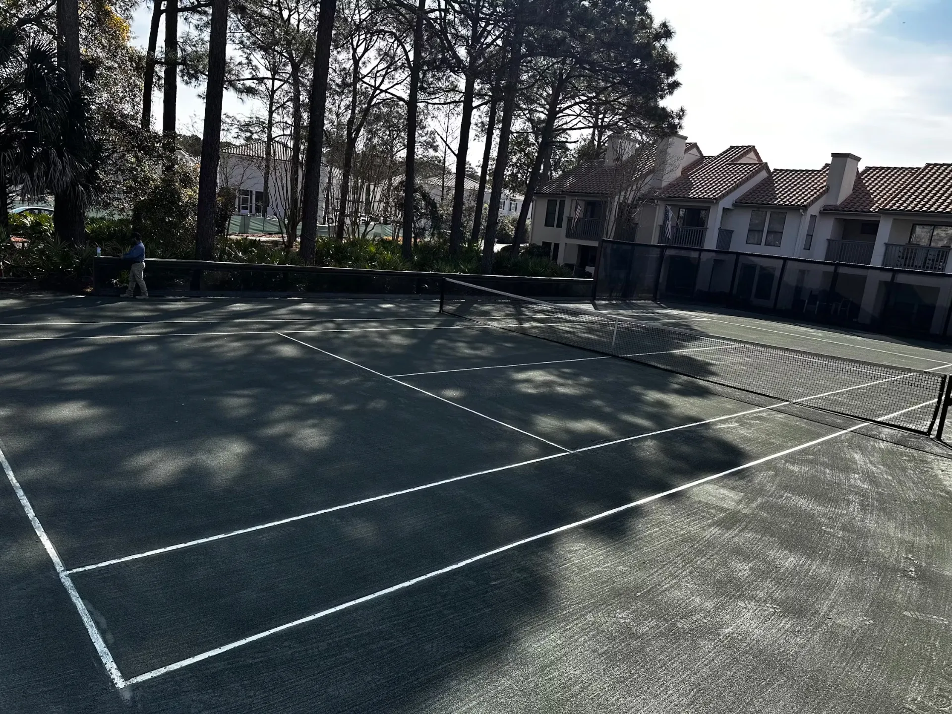 Tennis court with a net, white lines, and tree shadows beside residential-style buildings.