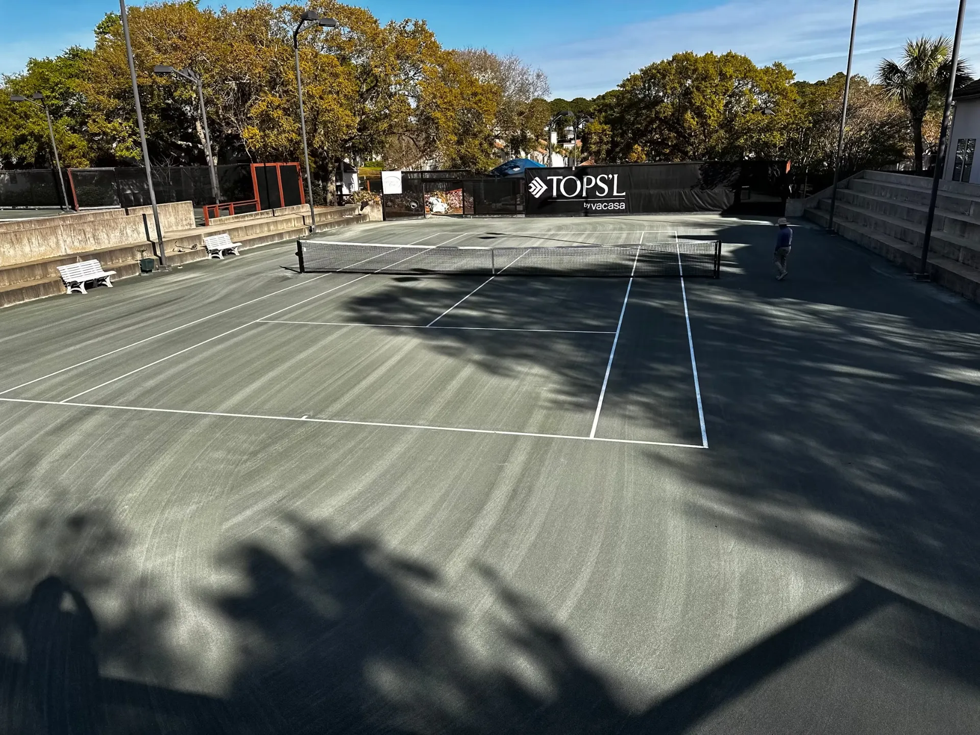 Tennis court with stepped seating and strong tree shadows across the surface.