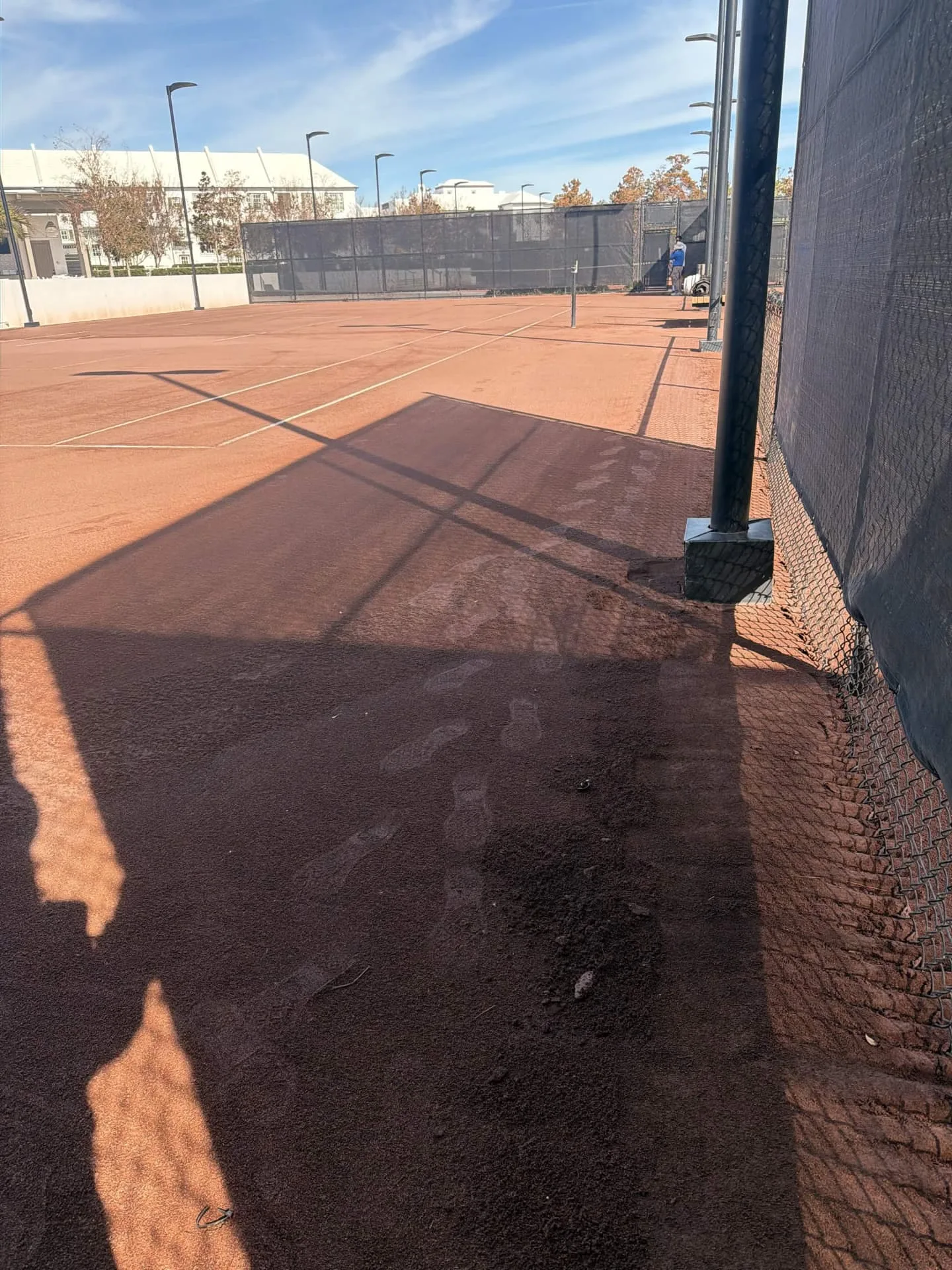 Wide view of a red clay tennis court before cleanup, with surface material tracks near the fence.