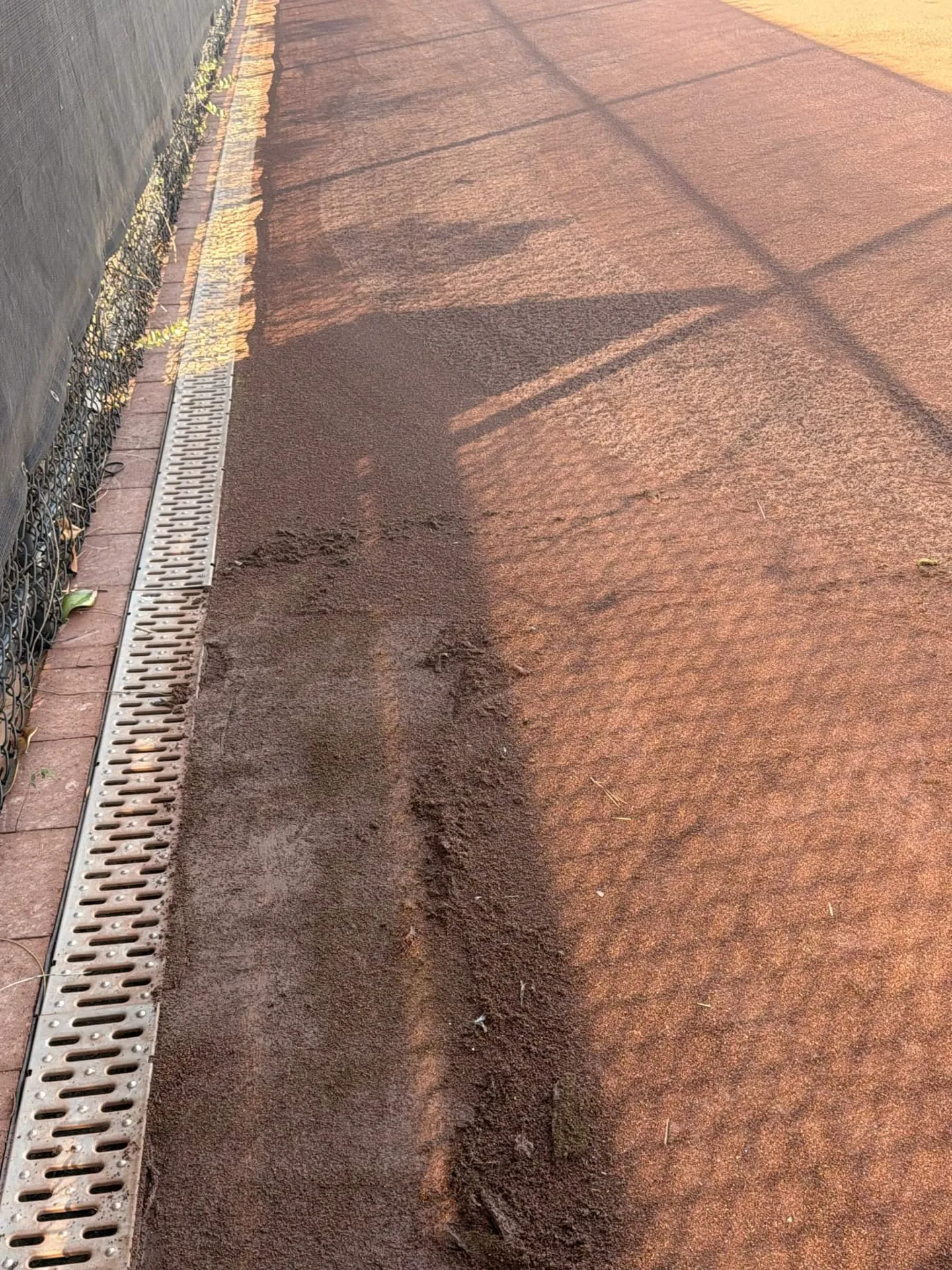 Loose clay surface material spread along the drainage strip at the edge of a court before cleanup.