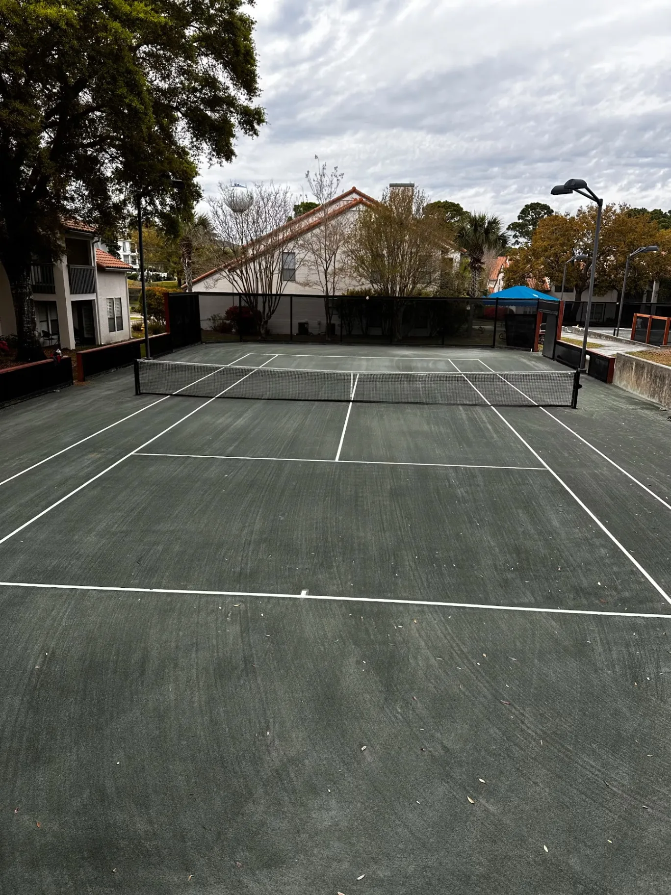 View along a tennis court toward a net, buildings, and trees.